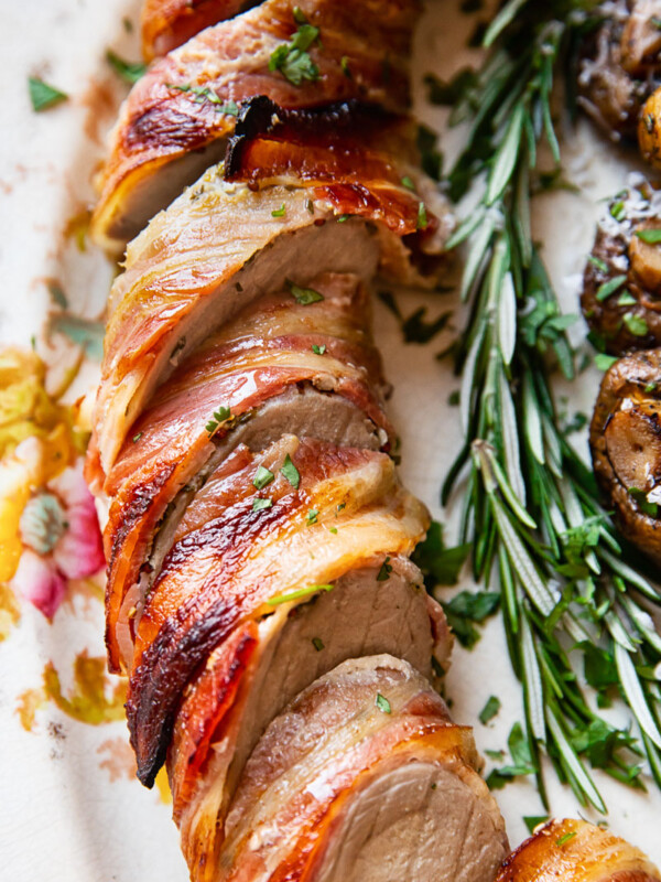 sliced pork tenderloin wrapped in bacon on a platter with rosemary sprig next to it
