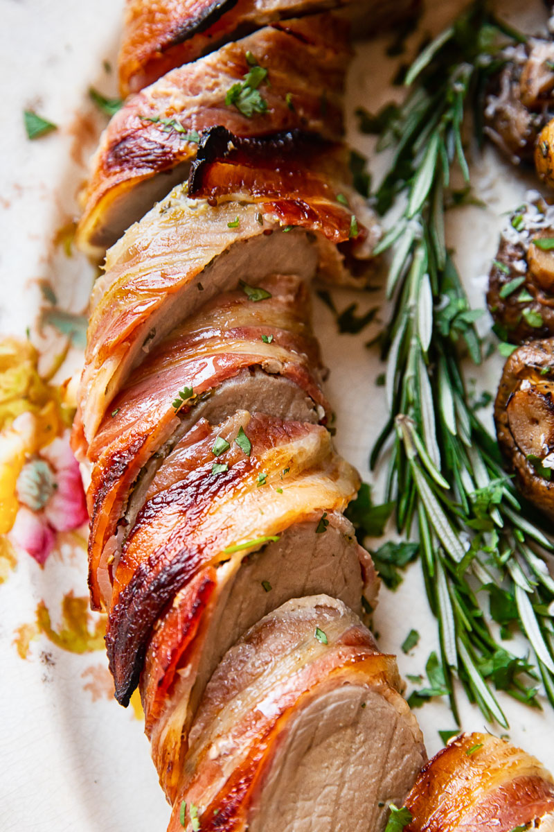 sliced pork tenderloin wrapped in bacon on a platter with rosemary sprig next to it