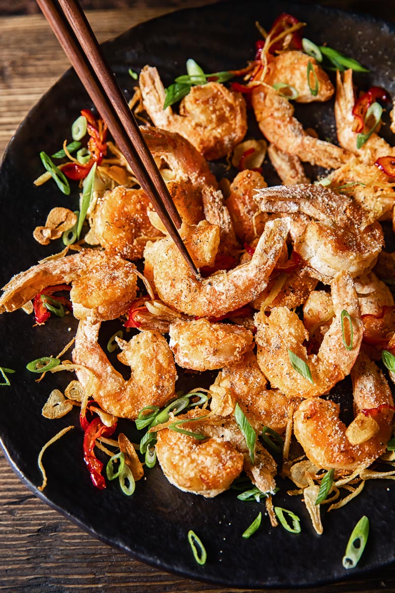 top down view of salt and pepper prawns on a dark platter, one shrimp being picked up by chopsticks