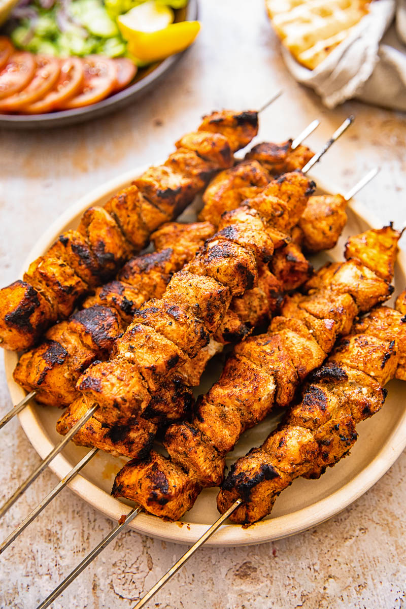 chicken shish kebab on a plate, cut up tomatoes and cucumbers in the background