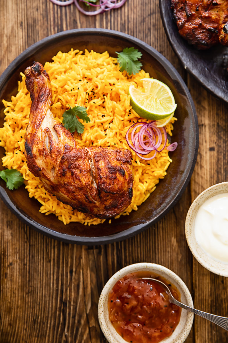 tandoori chicken on yellow rice on a brown plate, condiments around the plate