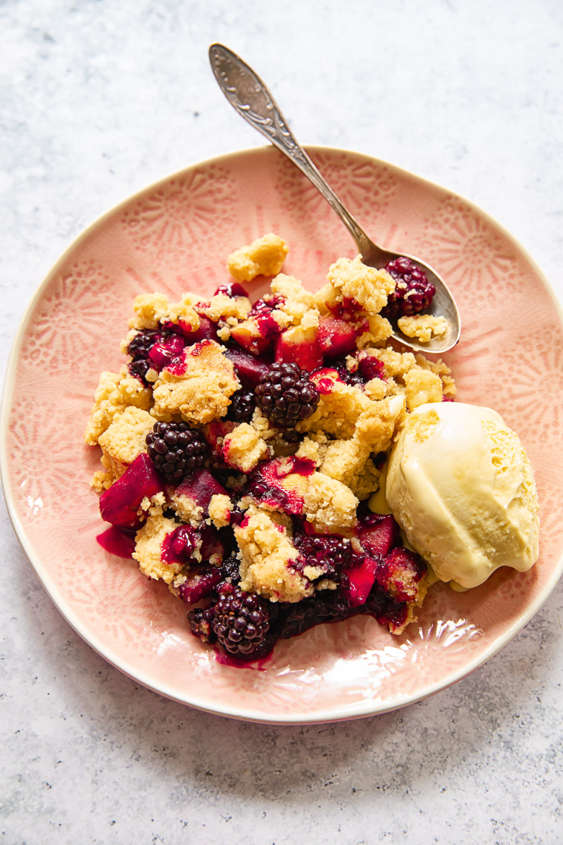 blackberry and apple crumble with a scoop of ice cream on a pink plate with a dessert spoon