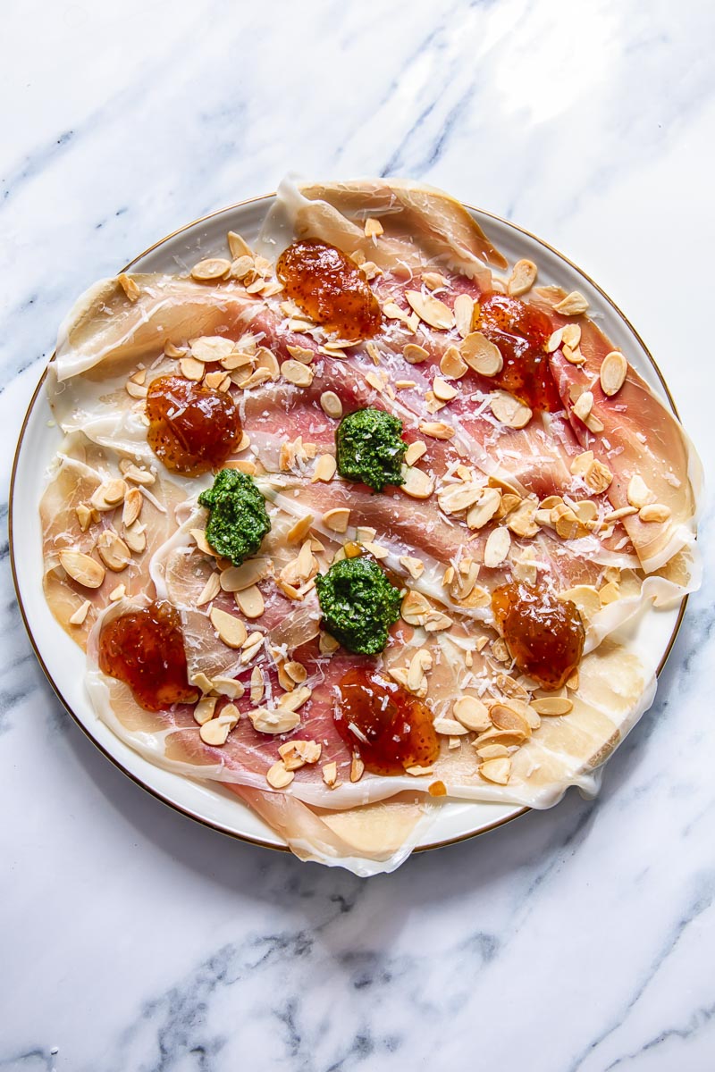 top down view of prosciutto platter topped with dollops of pesto, jam and almonds