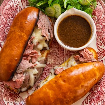 two long bread rolls filled with sliced toast beef and melted cheese with a dish of au jus on a platter