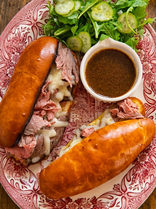 two long bread rolls filled with sliced toast beef and melted cheese with a dish of au jus on a platter