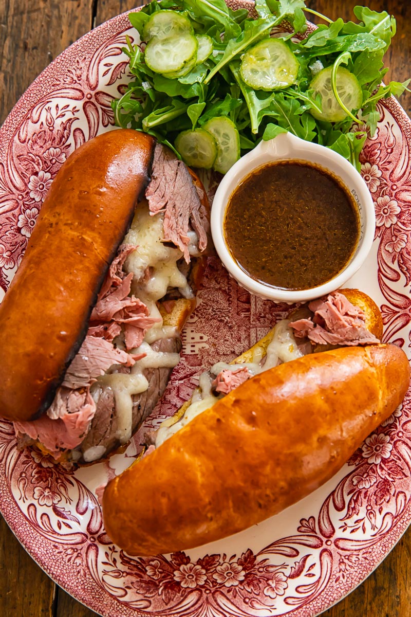 two long bread rolls filled with sliced toast beef and melted cheese with a dish of au jus on a platter