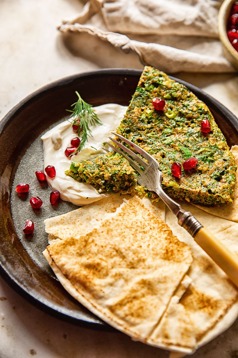 Kuku Sabzi on a brown plate on top of plain yogurt with flatbread and a fork