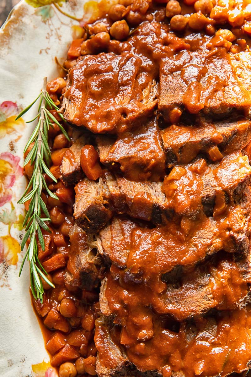 close up of sliced roast beef with a twig of fresh rosemary on the side in a rich sauce with chickpeas