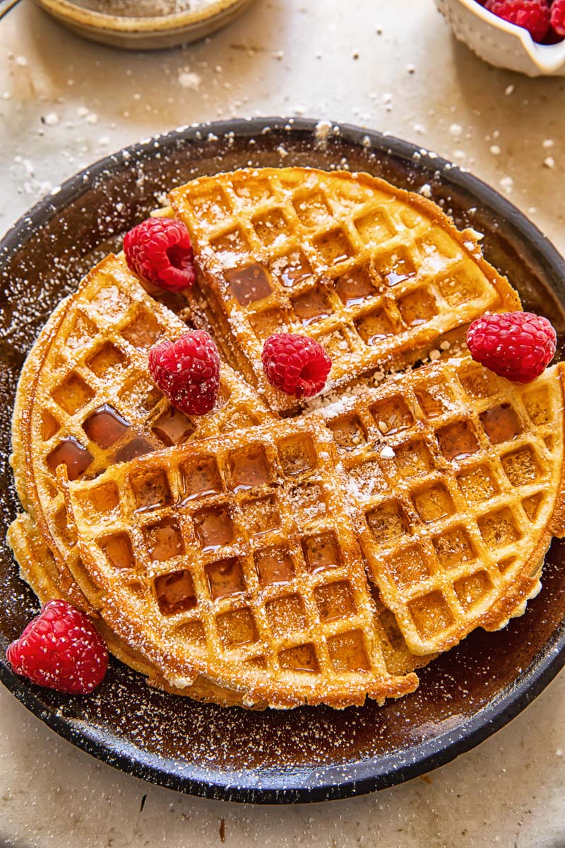 top down view of round waffles, topped with powdered sugar and raspberries on brown plate