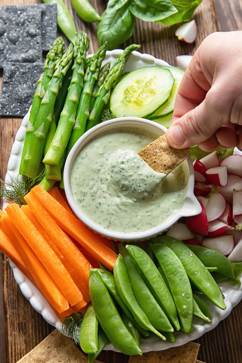 a cracker being dipped into green goddess dip and assorted vegetables on a platter