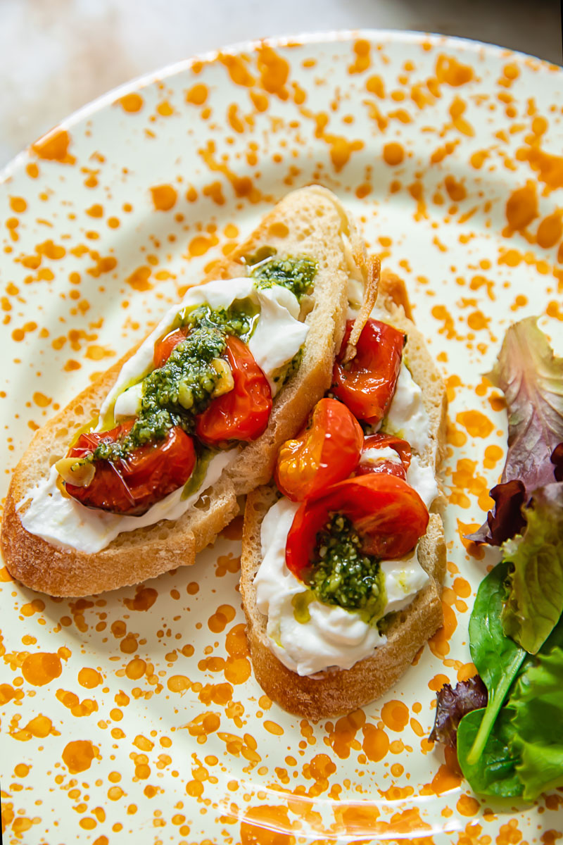 two slices of toast spread with ricotta, cherry tomatoes and pesto on orange speckled plate