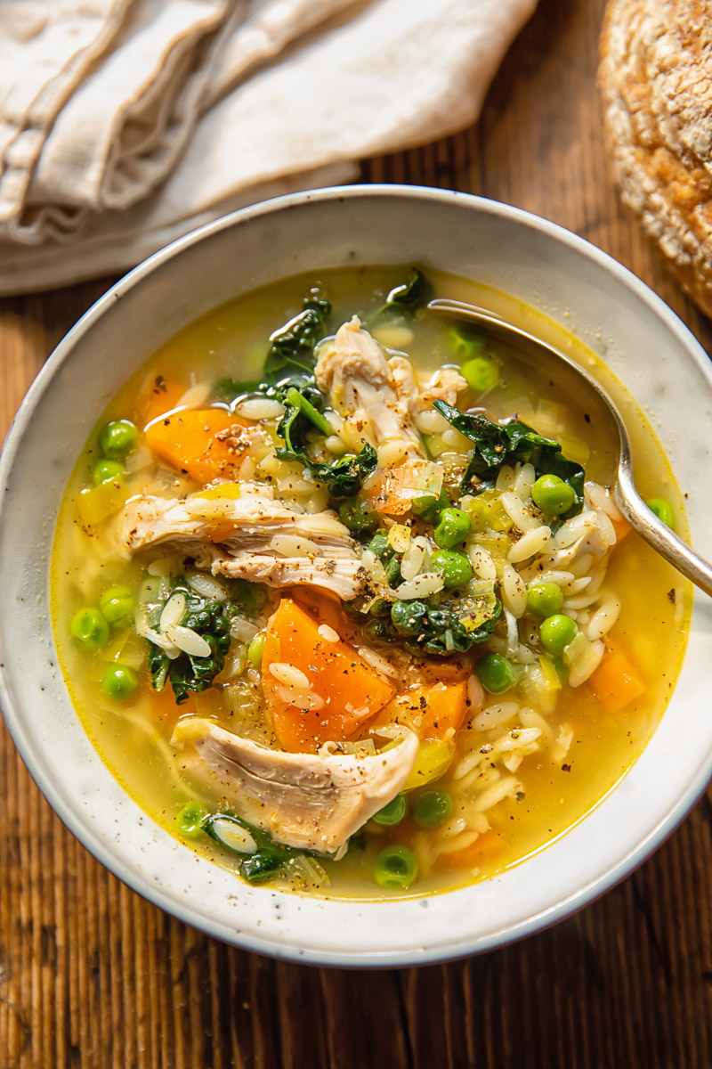 Top down view of chicken and vegetable soup with orzo, carrots and peas in a bowl with a spoon