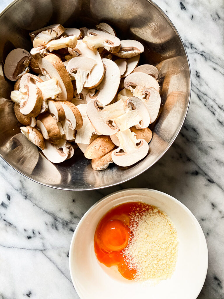 Top down of mushrooms in silver bowl next to bowl with one egg and cheese