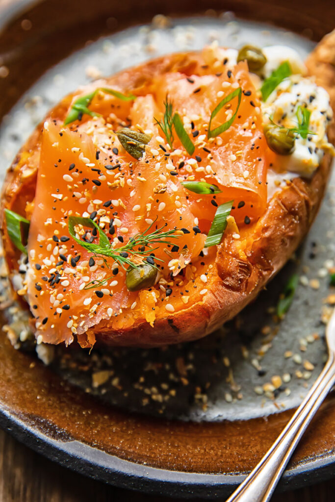 Close up shot of a baked sweet potato on a plate topped with sour cream, smoked salmon and capers