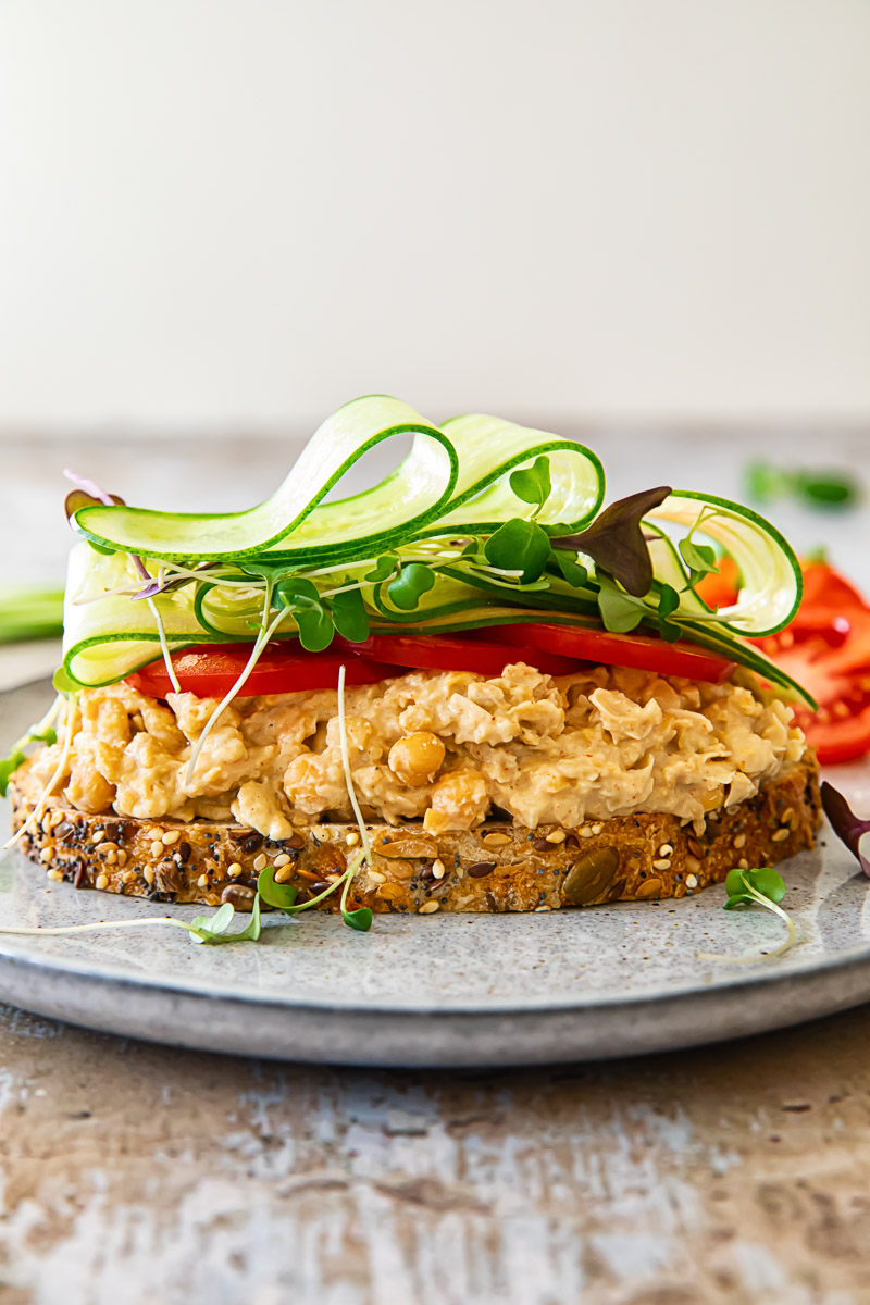 A chickpea salad sandwich with its top slice open, revealing layers of creamy chickpea salad, fresh tomatoes, and crisp microgreens on a plate.
