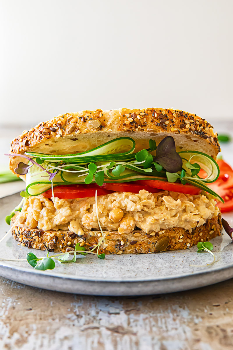 Front view of a chickpea salad sandwich, generously filled with salad and topped with heaping fresh vegetables, presented appealingly.
