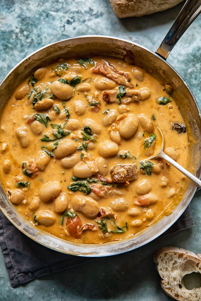 Top down shot of a spoon scooping tuscan butter beans in sun-dried tomato sauce with spinach out of a pan