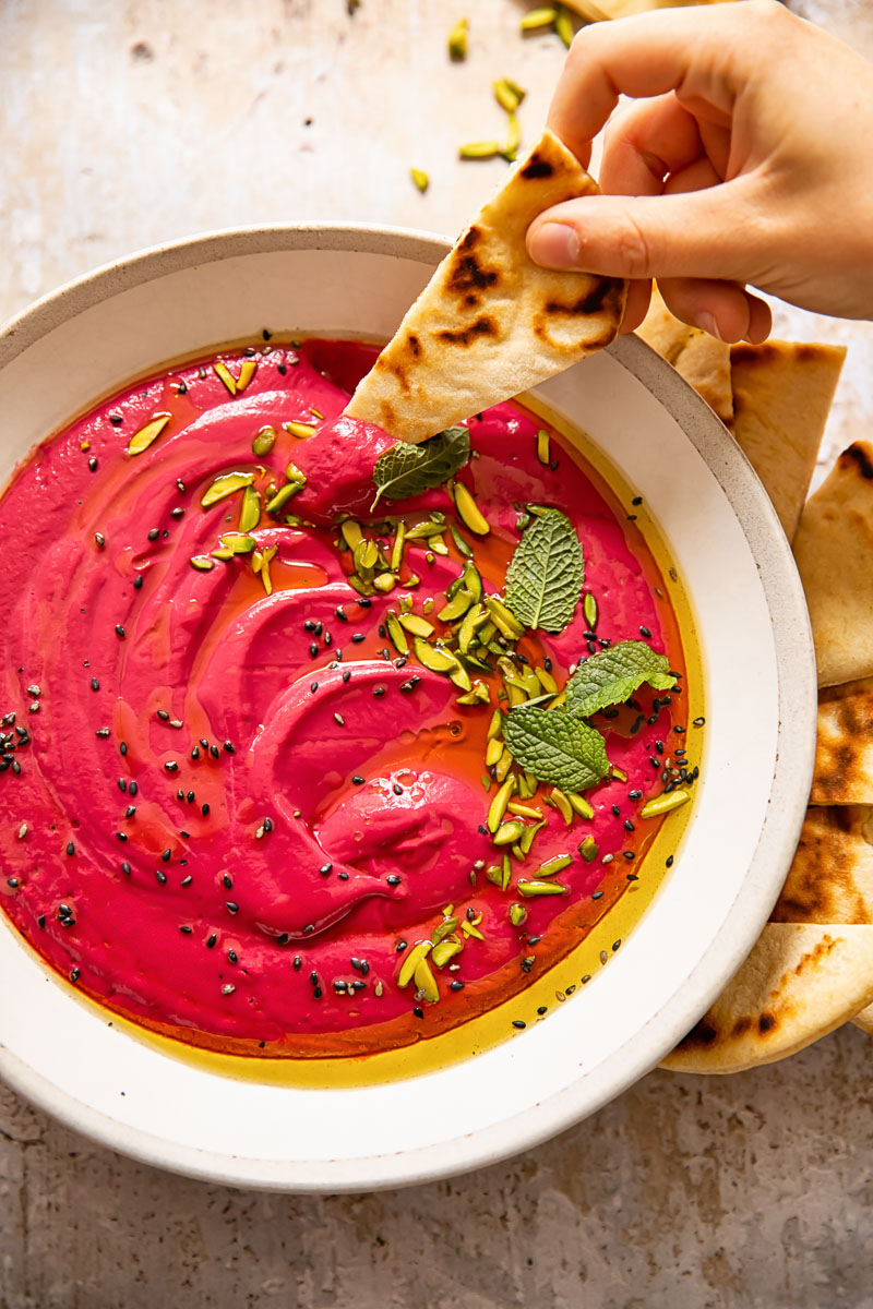 A hand dipping a piece of soft flatbread into a bowl of vivid pink beetroot borani dip, highlighting its creamy texture.