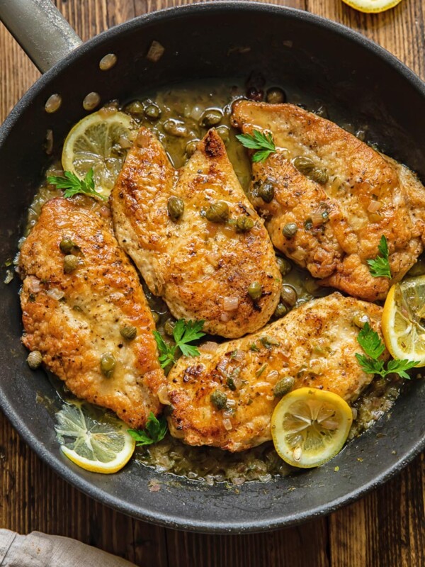 Top down view of chicken breasts in a pan, coated in piccata sauce with lemon slices and capers