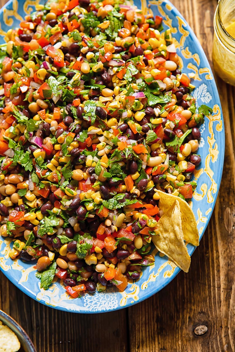 Top down shot of cowboy caviar on a large serving dish with beans, corn, pepper and tomatoes topped with dressing and fresh herbs