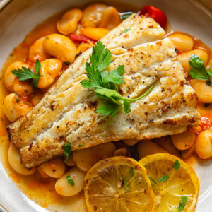 Close up shot of fried cod with lemon butter sauce in a bowl on top of crushed cherry tomatoes and butter beans