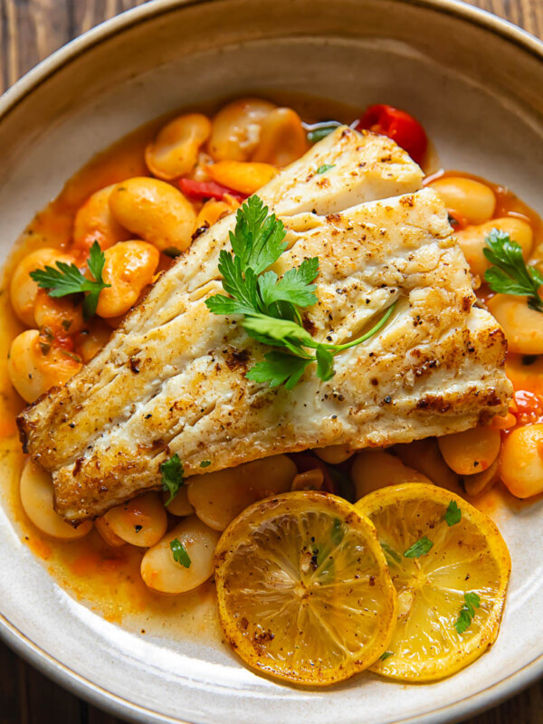 Close up shot of fried cod with lemon butter sauce in a bowl on top of crushed cherry tomatoes and butter beans
