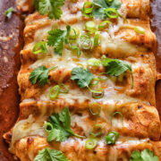 Close up shot of a row of chicken enchiladas in a baking dish topped with melted cheese and fresh herbs