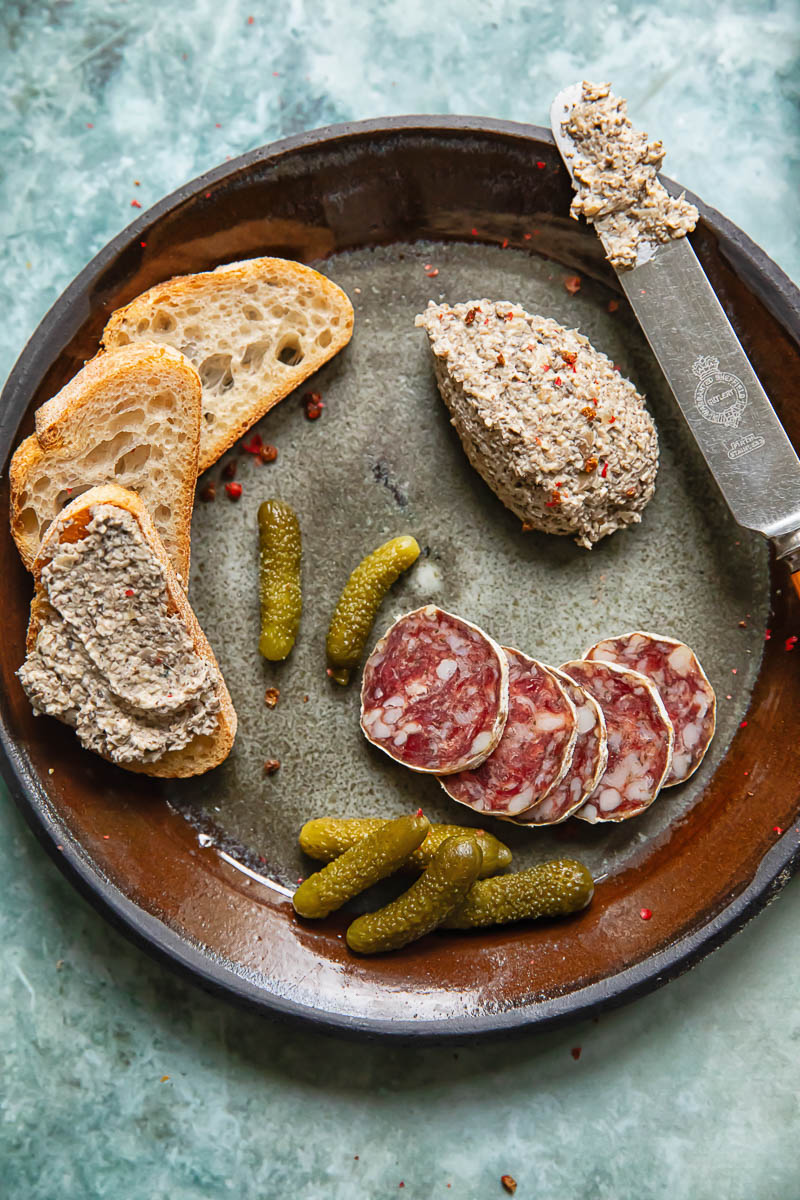 Shot of a beautifully arranged plate with mixed mushroom pâté alongside slices of artisanal bread, tangy gherkins, and savory salami slices, perfect for a gourmet spread.