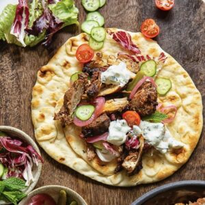 flatbread no a wooden background surrounded by vegetables, and topped with chicken, vegetables and tzatziki