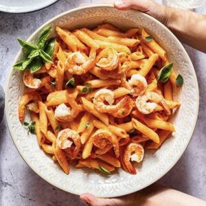 white bowl of penne pasta with shrimp, held by two hands