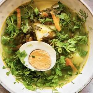 bowl of brothy soup with vibrant green vegetables and herbs, topped by a half a hardboiled egg