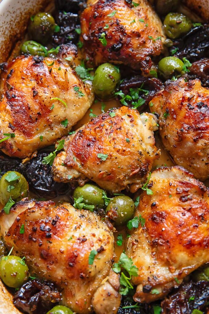 A close up of chicken thighs with a golden brown top, a sprinkling of herbs, alongside green olives and prunes