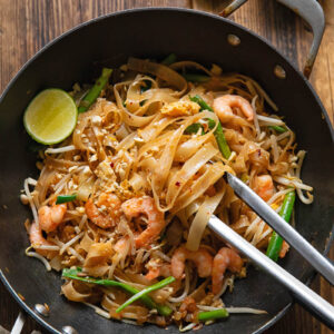 wok with pad Thai, with tongs removing a portion, next to a lime and peanuts