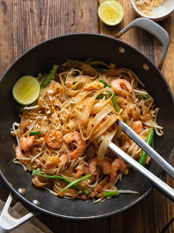 wok with pad Thai, with tongs removing a portion, next to a lime and peanuts