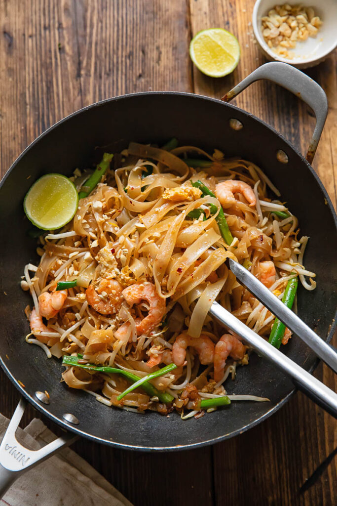 wok with pad Thai, with tongs removing a portion, next to a lime and peanuts