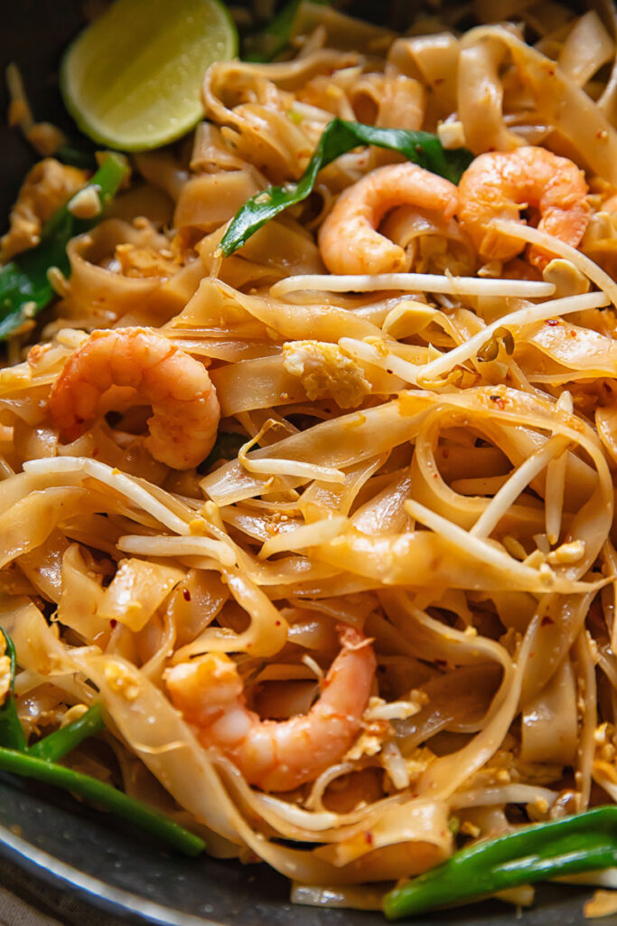 close up of noodles with shrimp, bean sprouts, green onions and a slice of lime