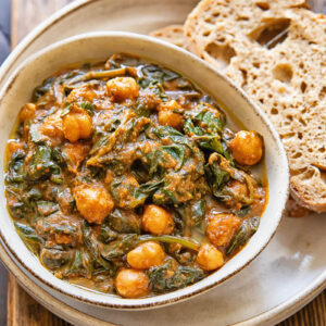 Top down view of a bowl of chickpeas and spinach, on top of a serving plate with a couple slices of bread.