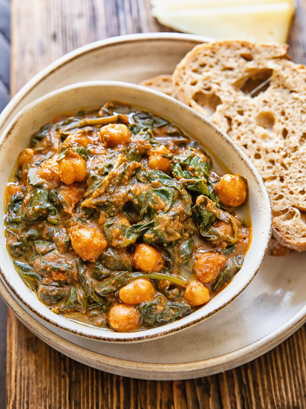 Top down view of a bowl of chickpeas and spinach, on top of a serving plate with a couple slices of bread.