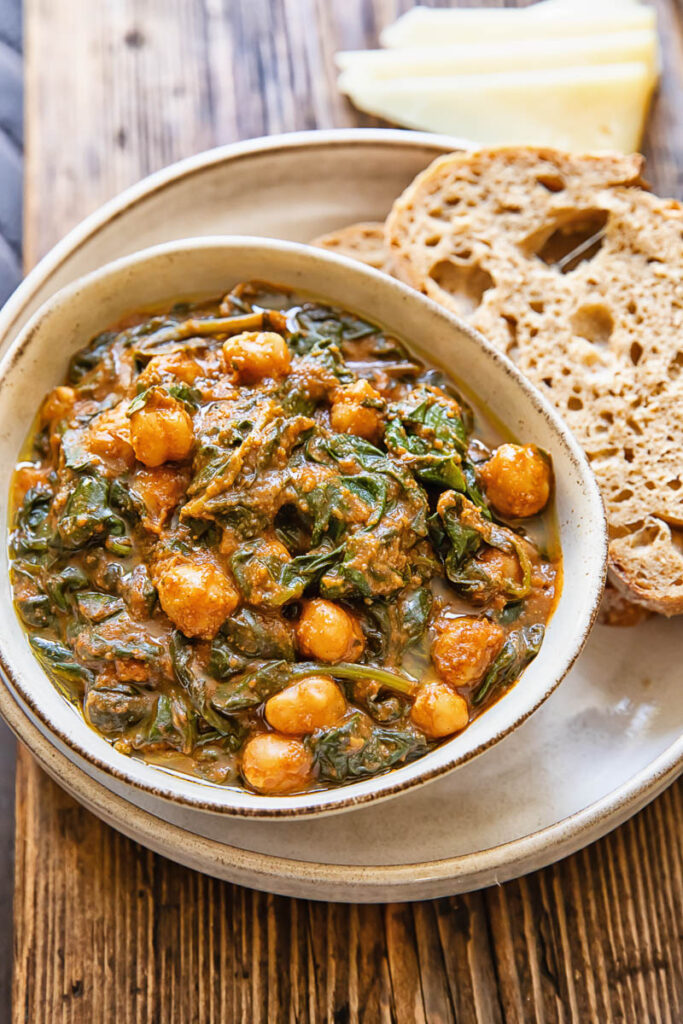 Top down view of a bowl of chickpeas and spinach, on top of a serving plate with a couple slices of bread.