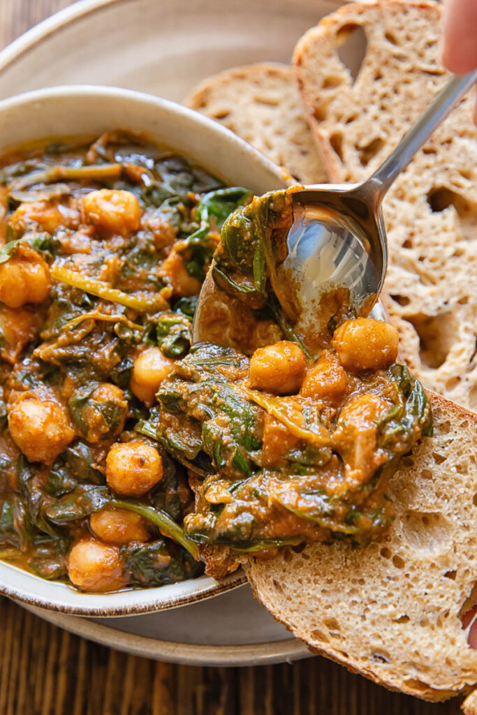 A spoon removing a portion of chickpeas and spinach from a bowl ot a pice of bread.