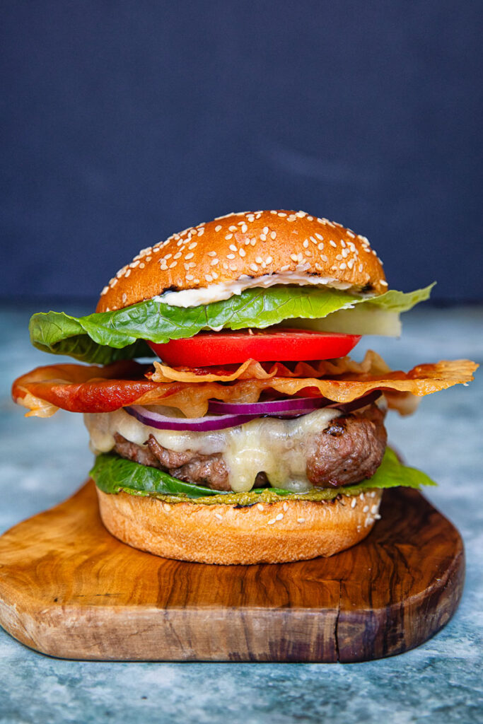 A vertical shot of a beef patty, tomato, lettuce and prosciutto in a bun against a dark background