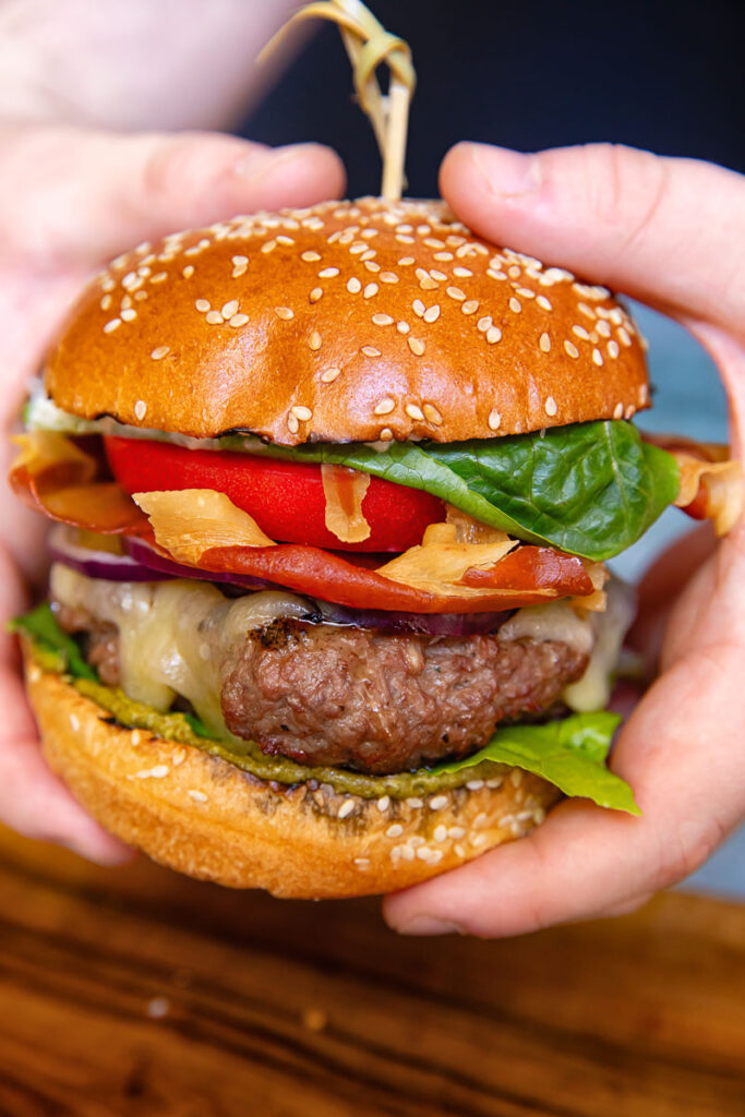 A vertical shot of a beef patty, tomato, lettuce and prosciutto in a bun in hands