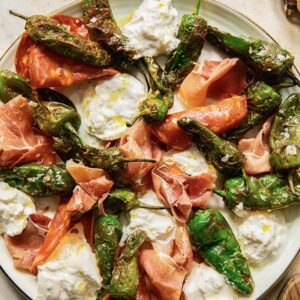 Square cropped photo of a white plate covered in chorizo slices, serrano ham, soft white cheese and fried padron peppersl