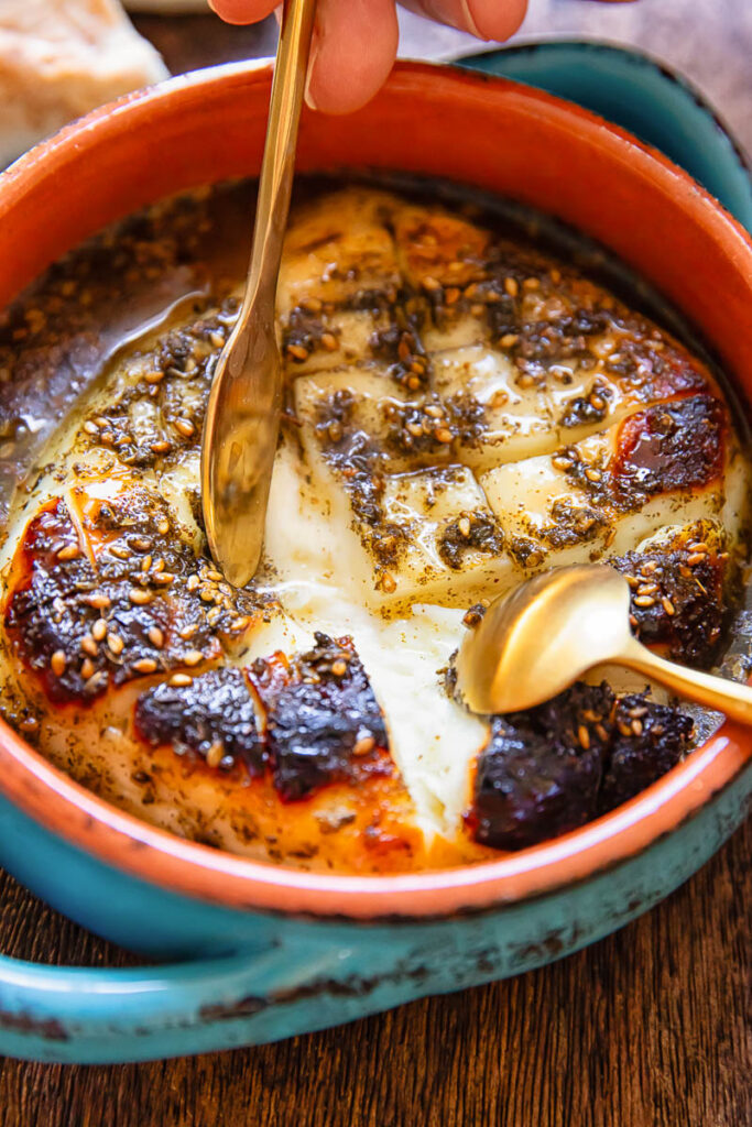 Close up of a small baking dish with baked halloumi being pulled apart by two small spoons.