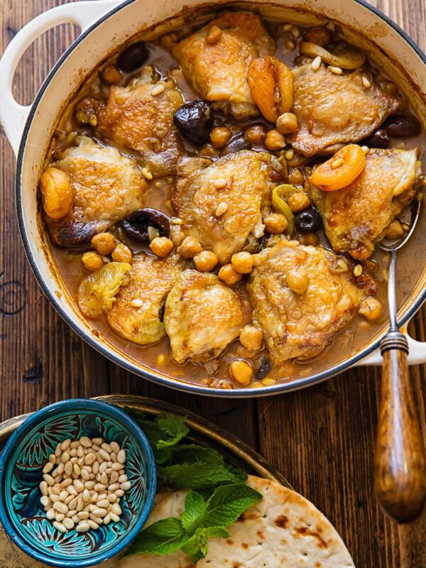 pan with chicken tagine on a wooden background next to a plate with flatbread and pine nuts