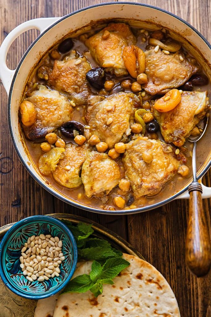 pan with chicken tagine on a wooden background next to a plate with flatbread and pine nuts