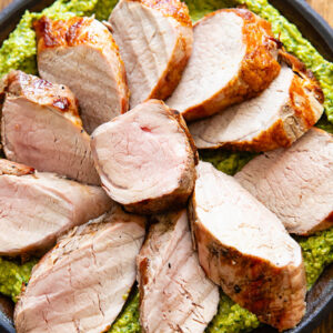Slices of pork tenderloin on a bed of green sauce, on a plate with a dark wood background.