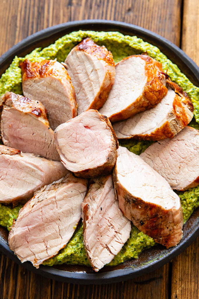 Slices of pork tenderloin on a bed of green sauce, on a plate with a dark wood background.