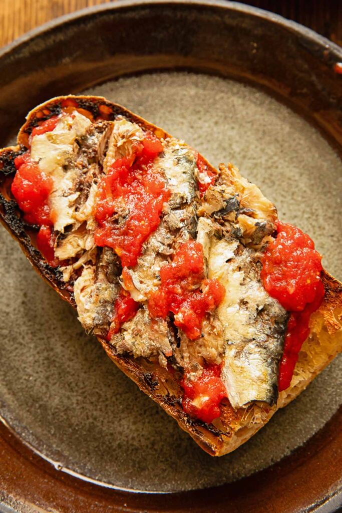 Toast on a dark plate with sardines and tomatoes.