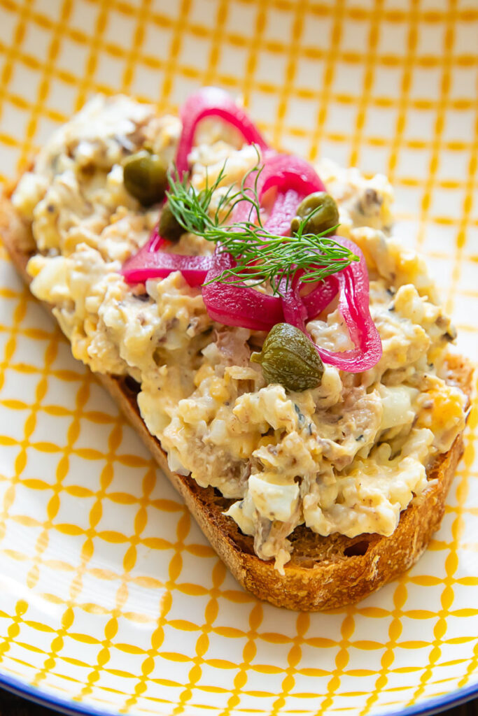Toast on a yellow striped plate topped by a sardine mixture, capers, pickled onion and dill.